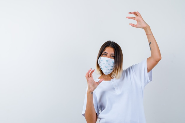 Junges Mädchen, das Hand streckt, als etwas imaginäres im weißen T-Shirt und in der Maske haltend und selbstbewusst aussehend. Vorderansicht.