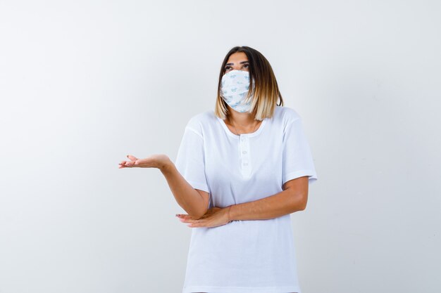 Junges Mädchen, das Handfläche beiseite spreizt, Hand unter Ellbogen im weißen T-Shirt, Maske hält und nachdenklich schaut. Vorderansicht.