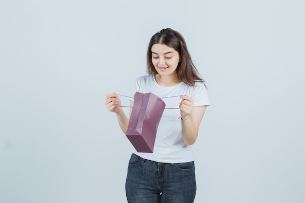 Junges Mädchen, das in eine Papiertüte in T-Shirt, Jeans schaut und glücklich schaut. Vorderansicht.