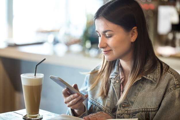 Junges Mädchen, das in einem Café sitzt, das Kaffee trinkt und das Telefon betrachtet