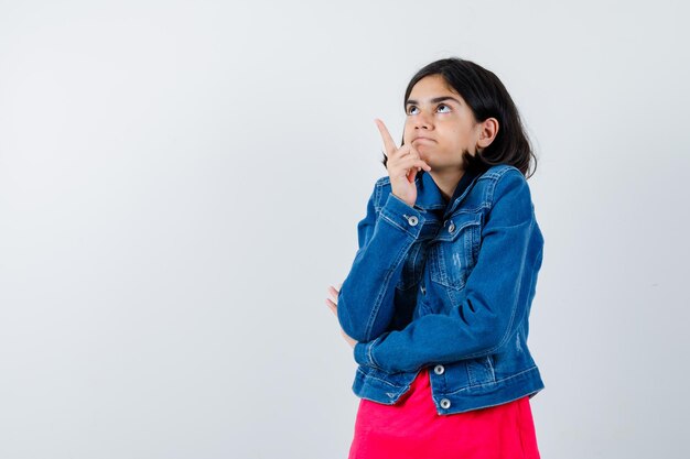 Junges Mädchen, das in rotem T-Shirt und Jeansjacke nach oben zeigt und glücklich aussieht, Vorderansicht.