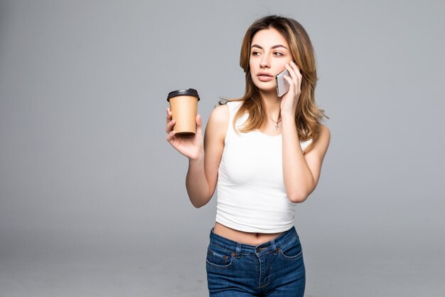 Junges Mädchen, das Kaffeetasse zum Mitnehmen hält, während auf Smartphone lokalisiert auf grauer Wand spricht