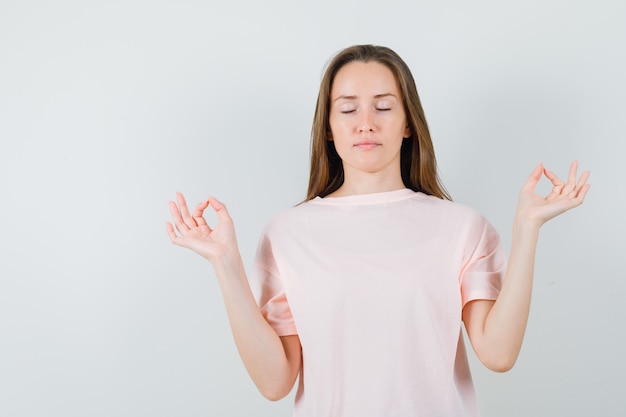 Junges Mädchen, das Meditation mit geschlossenen Augen im rosa T-Shirt tut und ruhig schaut. Vorderansicht.