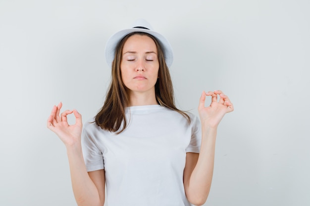 Junges Mädchen, das Meditation mit geschlossenen Augen im weißen T-Shirt, Hut tut und entspannt schaut, Vorderansicht.