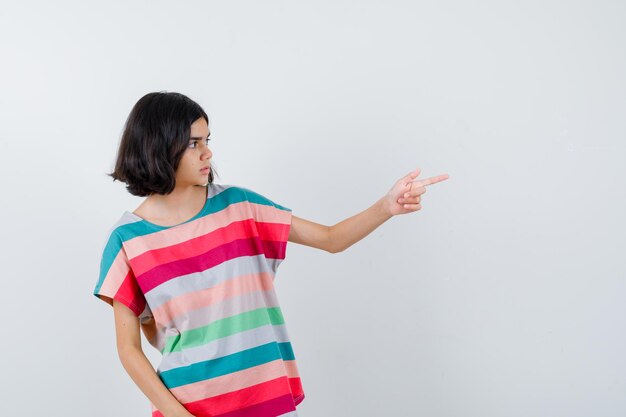 Junges Mädchen, das mit dem Zeigefinger im bunt gestreiften T-Shirt nach rechts zeigt und süß aussieht, Vorderansicht.