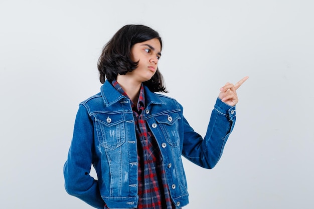 Junges Mädchen, das mit dem Zeigefinger in kariertem Hemd und Jeansjacke nach rechts zeigt und ruhig aussieht. Vorderansicht.