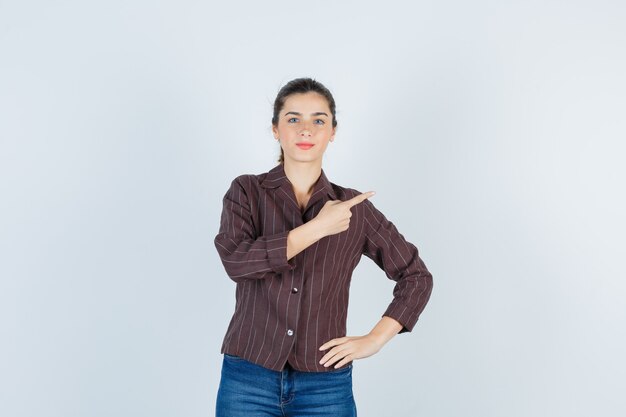 Junges Mädchen, das mit dem Zeigefinger nach oben zeigt, mit der Hand in der Hüfte in gestreiftem Hemd, Jeans und fröhlich aussehend, Vorderansicht.