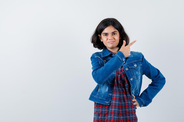 Junges Mädchen, das mit dem Zeigefinger nach rechts zeigt, während es die Hand in kariertem Hemd und Jeansjacke an der Taille hält und süß aussieht, Vorderansicht.