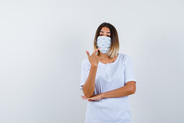 Junges Mädchen, das mit Zeigefinger auf sich selbst zeigt, Hand unter Ellbogen im weißen T-Shirt, Maske hält und ernst schaut, Vorderansicht.