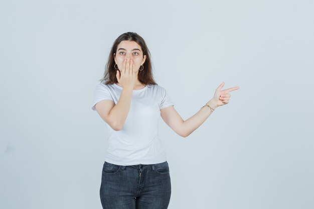 Junges Mädchen, das Mund mit einer Handfläche bedeckt, in T-Shirt, Jeans zur Seite zeigt und schockiert aussieht. Vorderansicht.