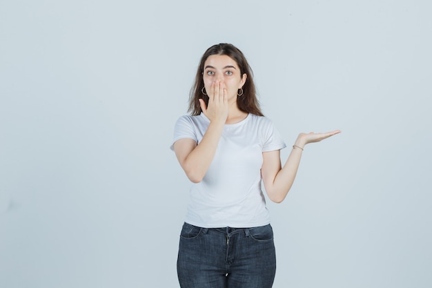 Junges Mädchen, das Mund mit Hand bedeckt, Handfläche in T-Shirt, Jeans beiseite spreizend und geschockt aussehend. Vorderansicht.