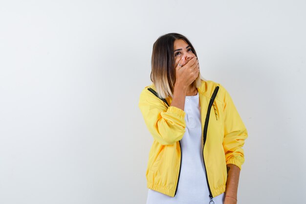 Junges Mädchen, das Mund mit Hand im weißen T-Shirt, gelbe Jacke bedeckt und fröhlich schaut, Vorderansicht.