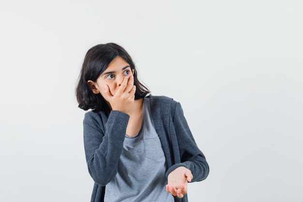 Junges Mädchen, das Mund mit Hand in hellgrauem T-Shirt und dunkelgrauem Kapuzenpullover mit Reißverschluss vorne bedeckt und überrascht aussieht.