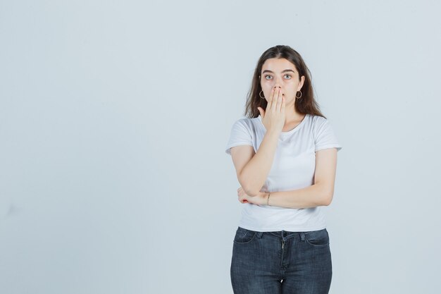 Junges Mädchen, das Mund mit Hand in T-Shirt, Jeans bedeckt und überrascht schaut. Vorderansicht.