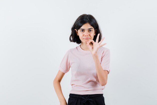 Junges Mädchen, das ok Zeichen im rosa T-Shirt und in der schwarzen Hose zeigt und glücklich schaut