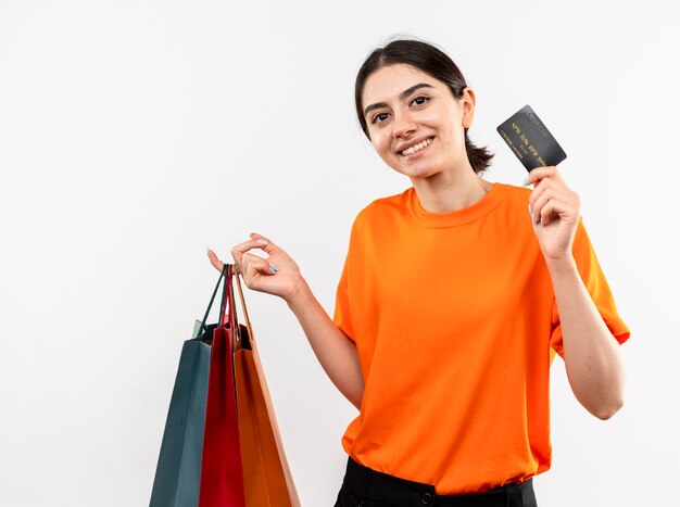 Junges Mädchen, das orange T-Shirt hält, das Papiertüten hält, die Kreditkarte lächelnd mit glücklichem Gesicht über weißer Wand stehen zeigt