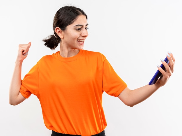 Junges Mädchen, das orange T-Shirt hält, das Smartphone geballte Faust glücklich und aufgeregt über weißer Wand hält