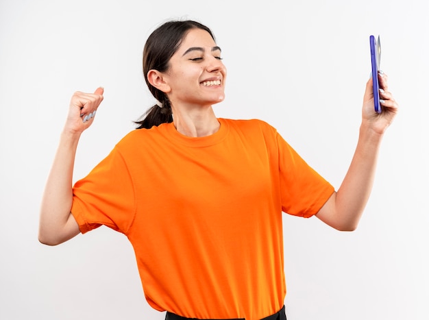 Junges Mädchen, das orange T-Shirt hält Smartphone, das Faust glücklich und aufgeregt über weißem Hintergrund zusammenhält