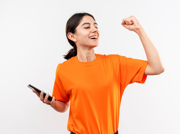 Junges Mädchen, das orange T-Shirt hält Smartphone hält geballte Faust glücklich und aufgeregt lächelnd fröhlich über ihren Erfolg stehend über weiße Wand