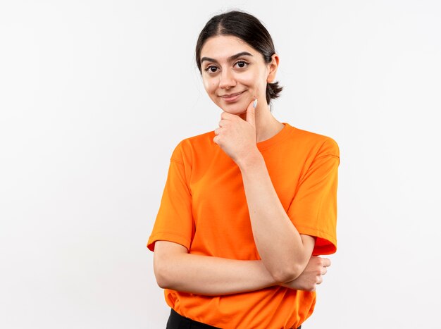 Junges Mädchen, das orange T-Shirt mit Hand auf Kinn denkend und lächelnd steht über weißer Wand trägt