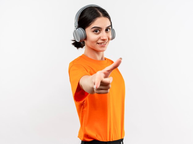 Junges Mädchen, das orange T-Shirt mit Kopfhörern glücklich und positiv genießt, die ihre Lieblingsmusik lächelnd mit Zeigefinger über weißer Wand stehend genießt