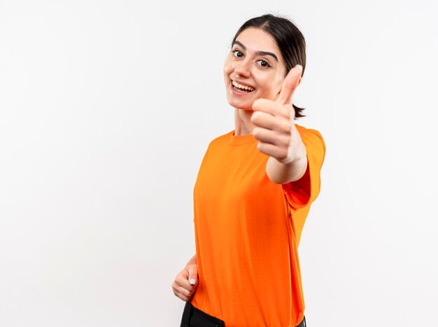 Junges Mädchen, das orange T-Shirt trägt, das die Kamera lächelt, die fröhlich zeigt Daumen hoch oben über weißem Hintergrund zeigt