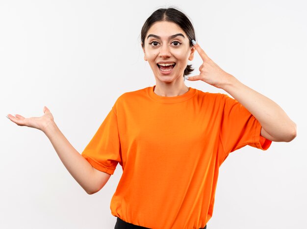 Junges Mädchen, das orange T-Shirt trägt, das etwas mit Arm zeigt, der Steinsymbol zeigt, das fröhlich über weißer Wand steht