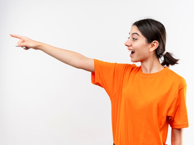 Junges Mädchen, das orange T-Shirt trägt, das mit Zeigefinger auf etwas lächelt, das mit glücklichem Gesicht über weißer Wand steht