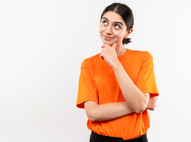 Junges Mädchen, das orange T-Shirt trägt, das oben mit Hand auf Kinn denkt und lächelt, das über weißer Wand steht