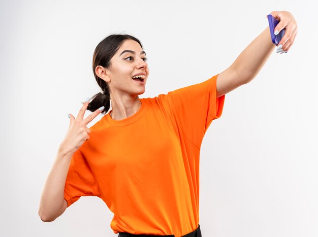 Junges Mädchen, das orange T-Shirt trägt, das Selfie unter Verwendung des lächelnden Smartphones tut, das V-Zeichen zeigt, das über weißer Wand steht