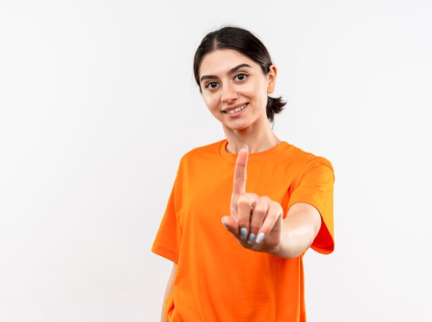 Junges Mädchen, das orange T-Shirt zeigt Zeigefinger mit Lächeln auf Gesicht steht über weißer Wand