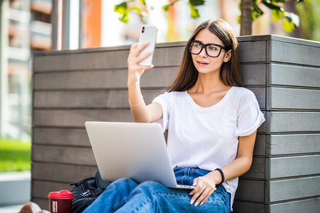 Junges Mädchen, das selfie auf Handy oder Videoanruf tut. Frau sitzt auf Bank, die an modernem Laptop-PC-Computer im Stadtpark in der Straße draußen auf Natur arbeitet. Freiberufliches Geschäftskonzept.