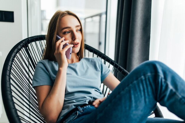 Junges Mädchen, das sich im Sessel ausruht und zu Hause in der Nähe des Fensters telefoniert?