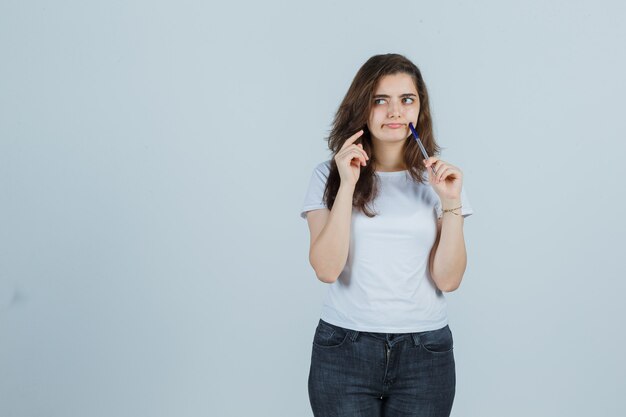 Junges Mädchen, das Stift auf Wange in T-Shirt, Jeans und nachdenklich aussehend, Vorderansicht hält.