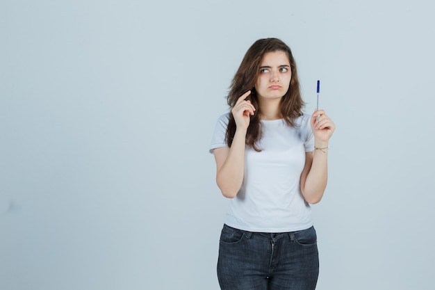 Junges Mädchen, das Stift hält, während in T-Shirt, Jeans denkend und nachdenklich schauend. Vorderansicht.