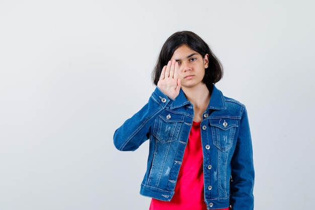 Junges Mädchen, das Stoppschild im roten T-Shirt und in der Jeansjacke zeigt und ernst schaut. Vorderansicht.