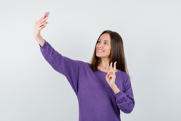 Junges Mädchen, das V-Zeichen zeigt, während Selfie im violetten Hemd nimmt und fröhlich schaut. Vorderansicht.