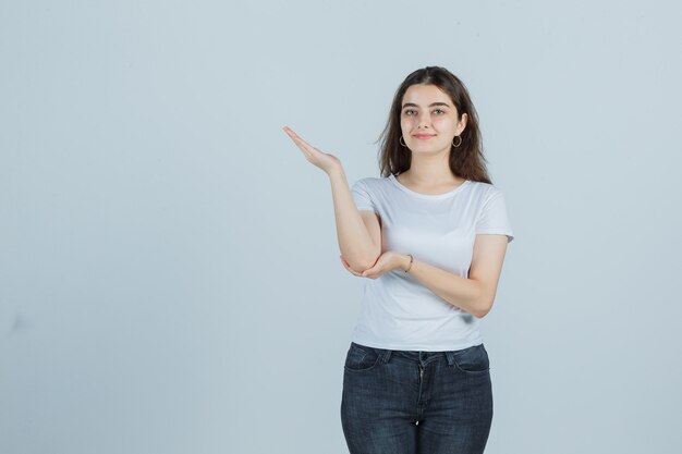 Junges Mädchen, das vorgibt, etwas im T-Shirt, in den Jeans zu zeigen und freudig zu sehen, Vorderansicht.