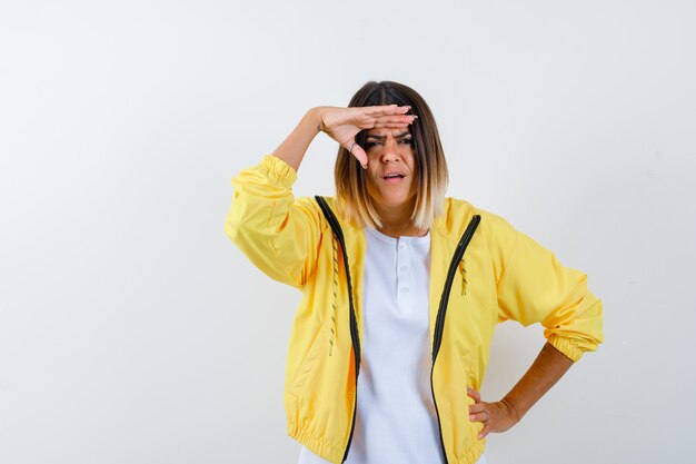 Junges Mädchen, das weit weg mit Hand über Kopf schaut, Hand auf Taille im weißen T-Shirt, gelbe Jacke setzt und konzentriert schaut. Vorderansicht.