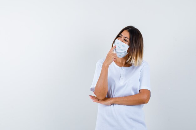 Junges Mädchen, das Zeigefinger auf Mund setzt, Hand unter Ellbogen in weißem T-Shirt und Maske hält und nachdenklich, Vorderansicht schaut.