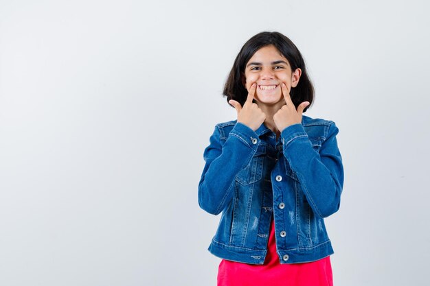 Junges Mädchen, das Zeigefinger in der Nähe des Mundes hält, ein Lächeln in rotem T-Shirt und Jeansjacke erzwingt und glücklich aussieht, Vorderansicht.