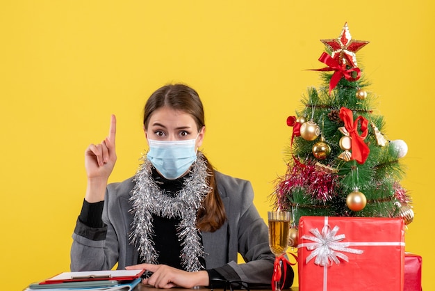 Junges Mädchen der Vorderansicht mit der medizinischen Maske, die am Tisch sitzt und etwas mit Fingerweihnachtsbaum und Geschenkcocktail zeigt