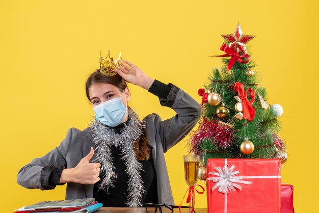 Junges Mädchen der Vorderansicht mit der medizinischen Maske, die Krone trägt, macht Daumen hoch Weihnachtsbaum und Geschenkcocktail