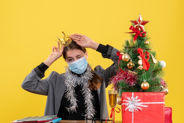 Junges Mädchen der Vorderansicht mit der medizinischen Maske, die Kronenweihnachtsbaum und Geschenkcocktail trägt