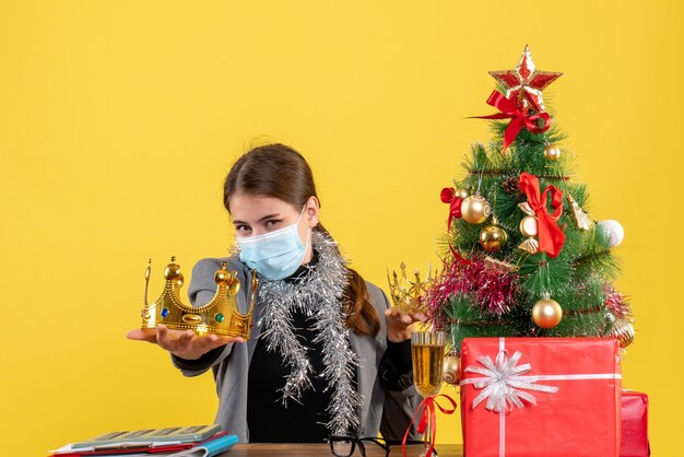 Junges Mädchen der Vorderansicht mit medizinischer Maske, die Kronenweihnachtsbaum und Geschenkcocktail zeigt