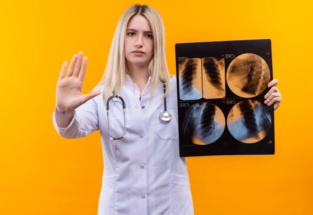 junges Mädchen des Arztes, das Stethoskop im medizinischen Kleid trägt, das Röntgenbild hält und Stoppgeste auf isolierter gelber Wand zeigt