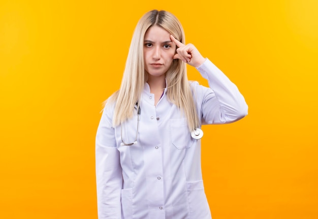 junges Mädchen des Arztes, das Stethoskop im medizinischen Kleid trägt, legte ihren Finger auf Stirn auf isolierte gelbe Wand