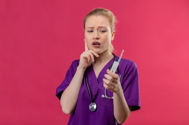 Junges Mädchen des denkenden Doktors, das lila medizinisches Kleid im Stethoskop hält, das Spritze hält, legte Hand auf Wange auf lokalisiertem rosa Hintergrund