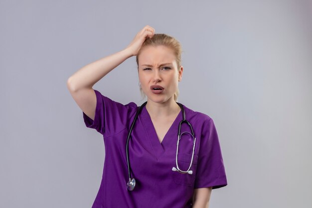 Junges Mädchen des denkenden Doktors, das lila medizinisches Kleid und Stethoskop trägt, ergriff Kopf auf lokalisiertem weißem Hintergrund