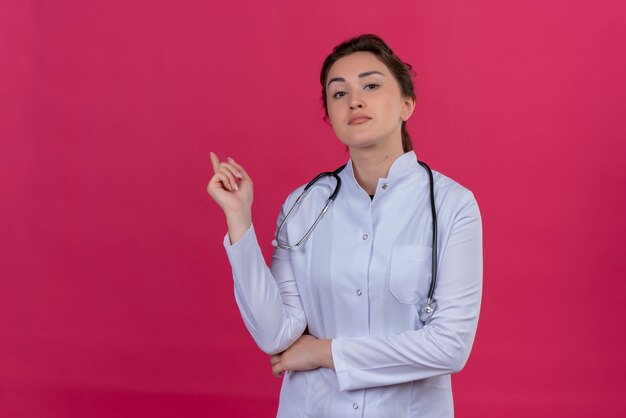 Junges Mädchen des denkenden Doktors, das medizinisches Kleid und Stethoskop trägt, zeigt auf isoliertem rotem Hintergrund zur Seite